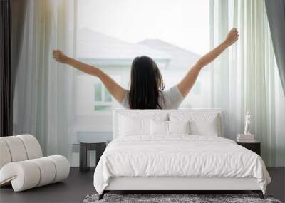 Rear view of Asian Woman stretching hands and body near window after wake up in bedroom at home. Concept for start new day with happiness. Young happy working female life Wall mural