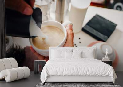 barista's hand making hot coffee mugs, latte art and pitcher made of milk froth, and there are coffee-making accessories on the white table. Close up Wall mural