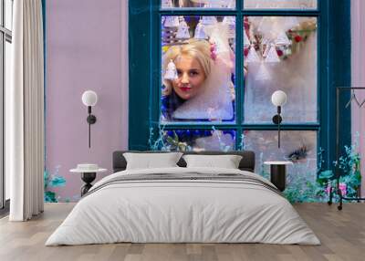 A young blonde woman looks through a window pane that is frozen and adorned with Christmas decorations. The concept of Christmas time. Wall mural