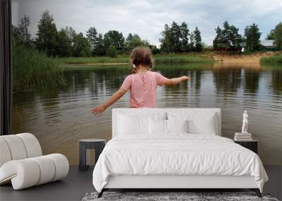 A little girl plays near the lake against the background of a country landscape. The concept of a happy childhood and outdoor recreation Wall mural