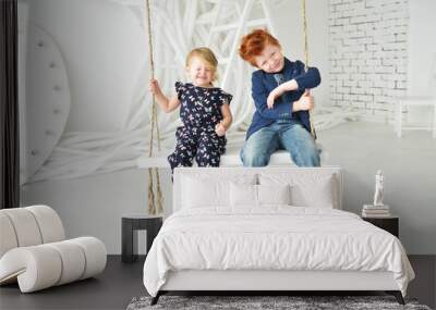 Cute kids red boy brother and blonde sister have a fun on the swing in the white studio Wall mural