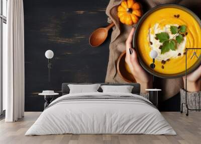 A woman s hands in a yellow sweater holding a pumpkin soup bowl on a dark stone background with a pumpkin decorated spoon Cozy autumn dinner idea from above Wall mural