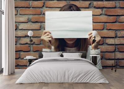 Caucasian woman holding white canvas, covering face, in front of brick wall. Wall mural