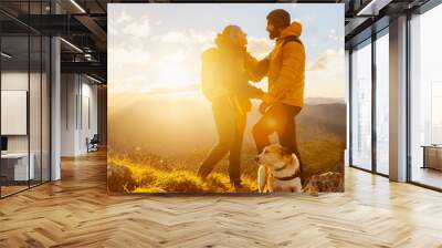 couple of hikers equipped with backpacks and warm clothing, standing on a mountain peak watching the sunset with their dog. outdoor sports and healthy lifestyle Wall mural