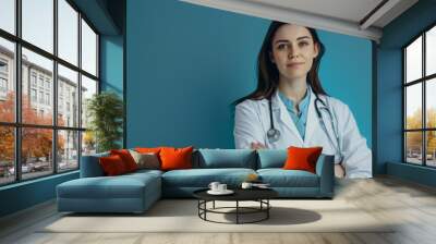 Confident young female doctor with glasses and stethoscope standing with crossed arms against a blue background. Wall mural