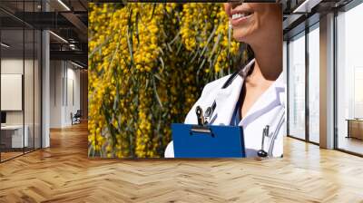 Unknown latina doctor woman smiles with a blue folder next to her chest as she goes to the daily review at the geriatric hospital where she works with a background of yellow flowers in the garden Wall mural