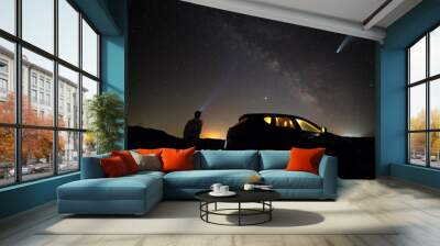 Silhouette of a man with head flashlight standing beside a car  lighting a beam of light on Milkyway galaxy and Neowise comet with light tail in dark night sky. Wall mural