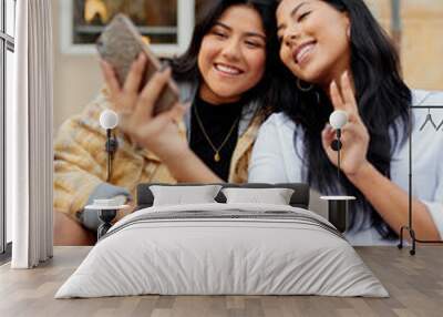 two hispanic sisters taking a picture with their smart phone in the street. Young latin girls using a mobile phone and having fun happily. Wall mural