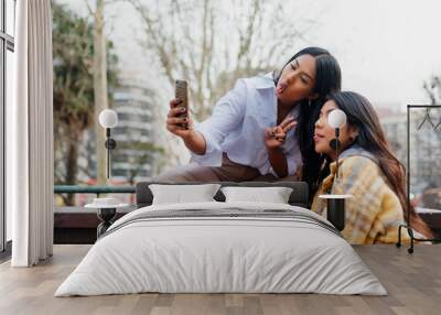 Two Latin girls sitting in the street taking pictures of themselves making faces with a phone. Sisters making a funny selfie for their family. Wall mural