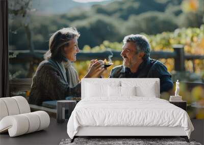 happy mid-aged couple having breakfast together at an outdoor table, the woman feeding the man food from her hand. The background is a sunny landscape of California wine country. Wall mural