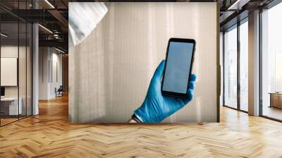 A man in a medical mask holds a smartphone with a white blank screen in his hand in a blue medical glove. Wall mural