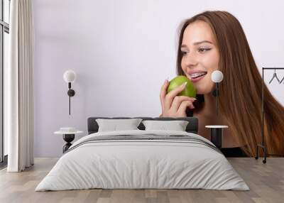 A young woman with braces on her teeth is smiling and eating a green apple. She is looking off to the side. The woman is wearing a black tank top and has long brown hair. Wall mural