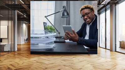 African American entrepreneur is sitting in a bright office at a desk by the window with a laptop and tablet. Black man with an Afro haircut looks into the camera and smiles while in the home office Wall mural