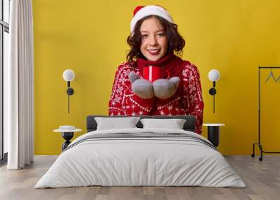 Beautiful woman with long brown hair wearing a red sweater adorned with snowflakes, a scarf, and a Santa hat. She is holding a red mug and smiling, set against a yellow background. Wall mural