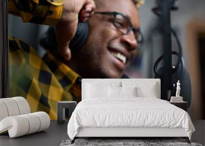 Close-up of an African podcaster blogger smiling while broadcasting his live audio podcast in the studio using a microphone and headphones. Male radio host with glasses, podcast host or interviewer Wall mural