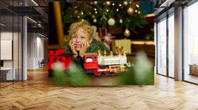 Happy curly-haired boy is playing with a magic toy train at the Christmas tree. The concept of celebrating New Year and Christmas. Wall mural