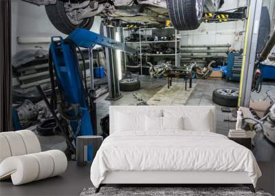 A raised car on a blue crane for engine or gearbox repair in a car workshop against the background of removed spare parts - wheels, rear axle axis. Wall mural