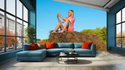 Blonde woman in bright pink suit and white T-shirt sits on haystack against background of mountains, trees and round rolls of hay with phone and takes selfie with hand outstretched. Summer vacation Wall mural