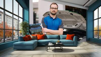 Portrait of handsome male mechanic hold auto repair tool. Man stand in front of car with open hood Wall mural