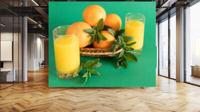  Two glasses of juice stand next to a wooden bowl filled with oranges and decorated with mint in soft natural light. Wall mural