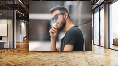 A young handsome white bearded man in black t-shirt smokes a cigarette in the street in the spring. Close up. Wall mural