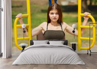 Young athletic caucasian girl in a striped top doing exercises on the simulator on the street sports field on a sunny day in summer. Fitness on street sports ground. Wall mural