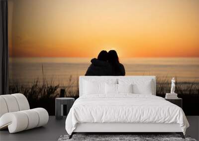 Silhouette of two female friends enjoying a conversation while sitting hugging and watching the sunset on a beach. Back view Wall mural