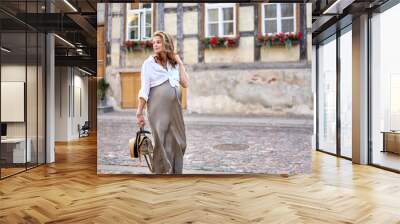 Young pregnant woman in with long curly hairstyle holding bag in hand is walking at the old sunny street of an old town. Wall mural