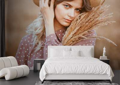 A young girl with long curly hair and a hat , in a brown dress with white polka dots, stands in a wheat field Wall mural