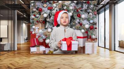 Cute happy child in santa hat holding a surprise gift on the background of a decorated Christmas tree Wall mural