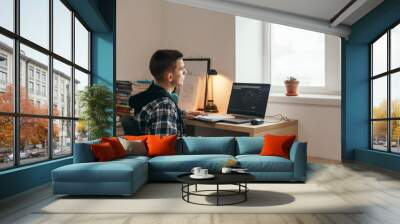 Teenage boy doing homework using computer sitting by desk in room alone Wall mural