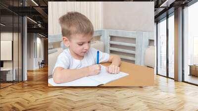 The child studies online at home. Cute little boy writes homework in a notebook sitting at the table at home. Online home education concept Wall mural