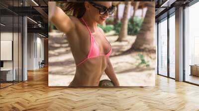 Young beautiful fit sporty female model pose in micro pink tight bikini at the beach with coconut in her hands Wall mural