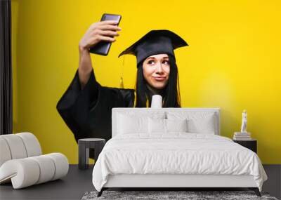 graduate woman makes selfie photo on phone over yellow background, concept of successful completion of studies, Wall mural