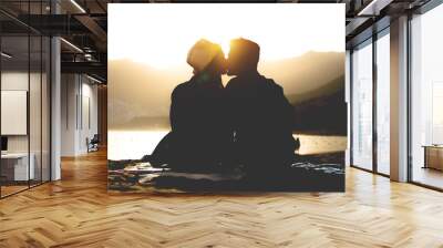 Romantic young couple kissing on the beach on sunset - Silhouette of teens lovers at the beginning of their story sitting on sand - People, love, lifestyle, relationship concept Wall mural