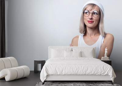 Cute blond young woman in glasses, a white hat looks to the side on a light background Wall mural