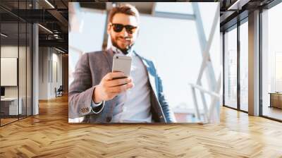 Young handsome businessman with stubble and sunglasses walking down the stairs outside. Smiling and looking at mobile phone Wall mural