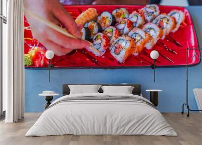 Hand using chopsticks to pick up sushi from colorful platter with assorted sushi rolls and heart-shaped soy sauce dish. Curacao. Wall mural