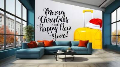 On a yellow plastic suitcase wearing a red Santa Claus hat on a light background. Added the inscription Merry Christmas and Happy New Year Wall mural