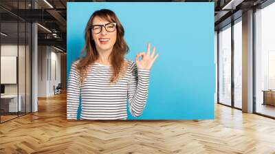 Portrait of a young friendly woman with a smile in a casual t-shirt and glasses makes an okay gesture on an isolated light background. Emotional face. Gesture is all right, it's OK Wall mural