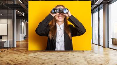 smiling young business woman looking through binoculars on yellow background Wall mural