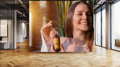 Happy young lovely woman holding massage oil with pipette, smiling looking away. Pretty joyful lady with cosmetic oil in hands at window sunshine. Cosmetology self care concept. Copy ad text space Wall mural