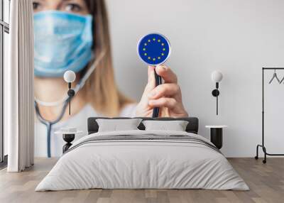 young woman in a medical mask holds a stethoscope with the flag of european union on a light backgro Wall mural