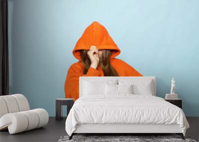 Young woman in orange hoodie on blue background hides face under hood Wall mural