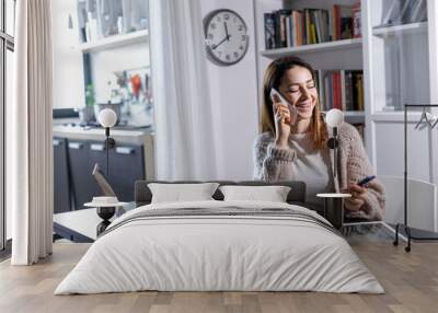 Giovane donna pratica lo smart working da casa, e parla al telefono sorridendo , sullo sfondo la cucina Wall mural