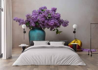 A large bouquet of lilacs in a vase on a light gray background, next to them on a plate are bananas, two red apples, and an orange. Still life, plenty of space for text Wall mural