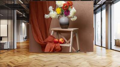 A large bouquet of white, yellow, red, autumn flowers. Many asters in a gray vase on a wooden staircase. Below are two red apples, a small pumpkin. Orange background. Orange curtain. Still life Wall mural