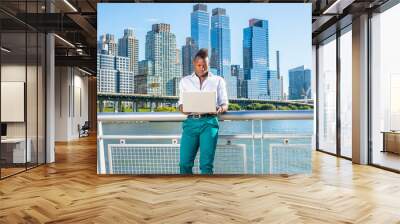 Dressing in a white shirt and green pants,  a young black guy with mohawk hair is standing in the front of high buildings by the water, working on a computer.. Wall mural