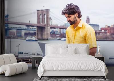 Raining - grainy, drizzling, wet feel. Young Man with beard traveling in New York, wearing short sleeve yellow shirt, sitting by East River, working on laptop computer. Bridges, boat on background.. Wall mural