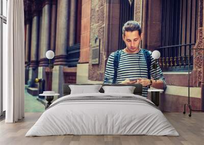 Russian Man traveling in New York, wearing striped long sleeve T shirt, shoulder carrying back bag, standing by wall on vintage street, reading, texting on cell phone, listening music with earphone.. Wall mural